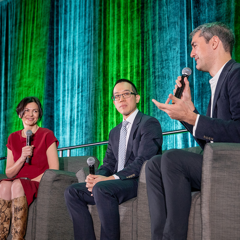 Three individuals seated on a stage during a panel discussion, each holding a microphone, with a backdrop of green and blue curtains.